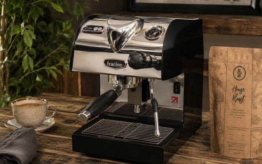 Luxury Fracino home espresso machine styled on a rustic wooden coffee bar with polished chrome finish, BE AR House Roast speciality coffee bag, ceramic espresso cup, soft natural lighting, indoor plants and warm artisan café atmosphere