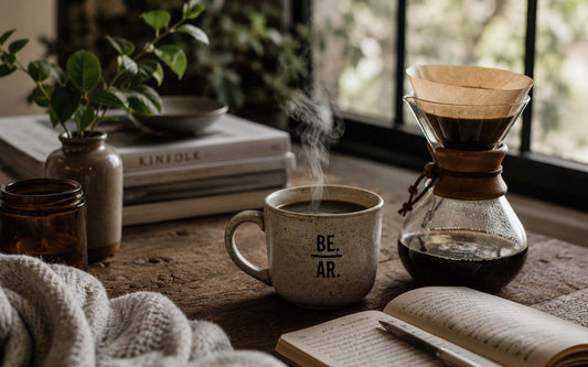 Freshly brewed pour-over coffee and a steaming ceramic mug on a rustic wooden table beside an open notebook, books, and houseplants, creating a calm morning coffee ritual in natural window light.