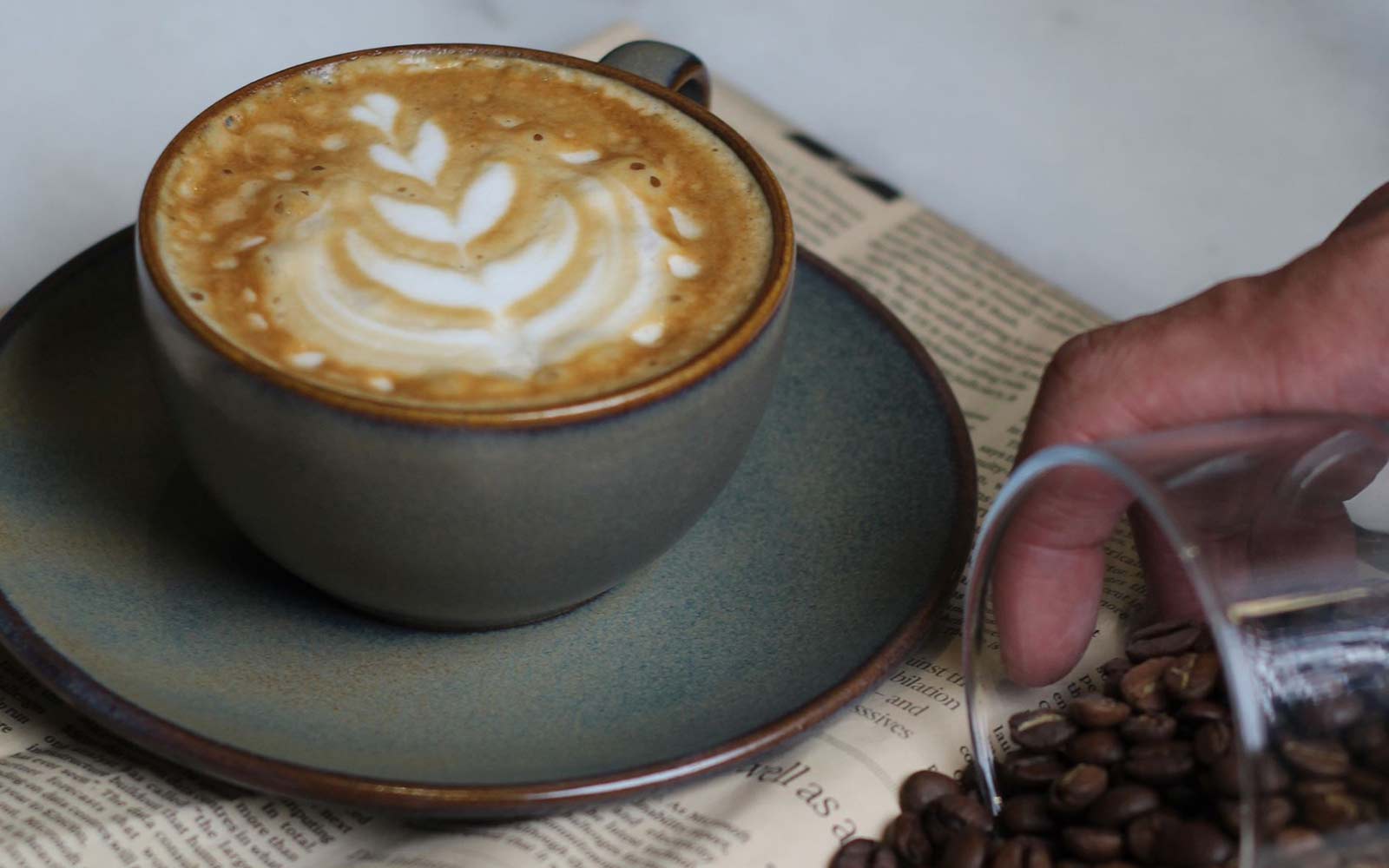 photo of a coffee with latte art of a leaf on top, hand pouring arabica coffee beans all over a book